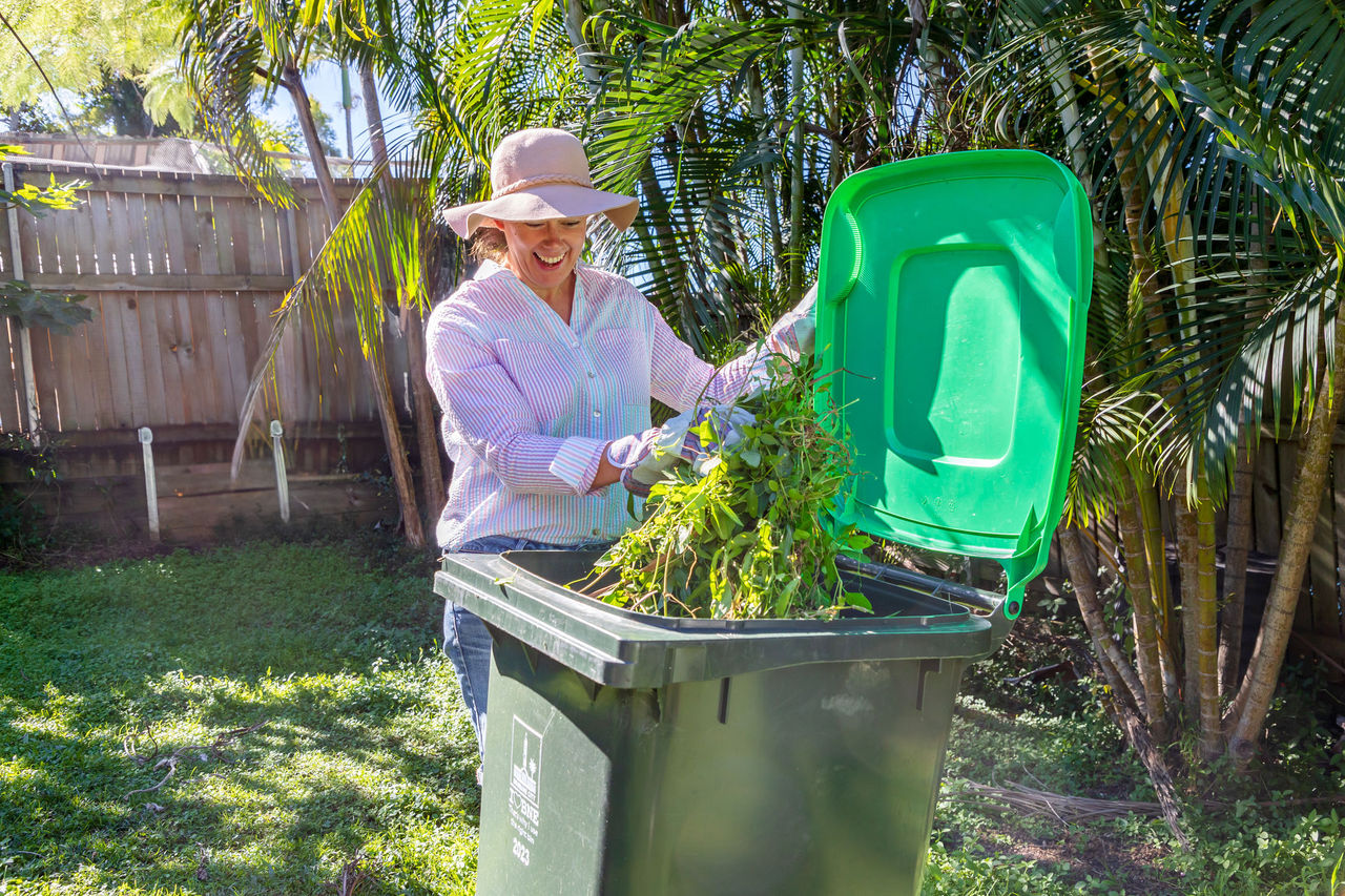 Woman disposing of vegetation in a green lidded wheelie bin.