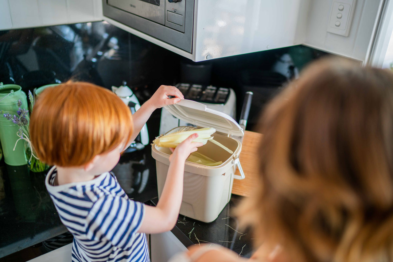 Young boy placing organic waste (appears to be a corn husk)in a kitchen benchtop caddy. Boy's Mum oversees him to his right.
