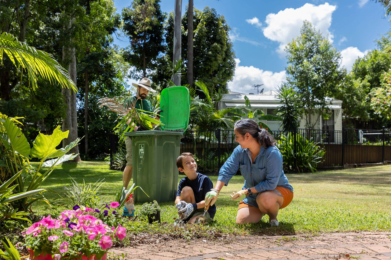 A man, woman and young boy gardening in a backyard with the man placing palm fronds into a green lidded bin.