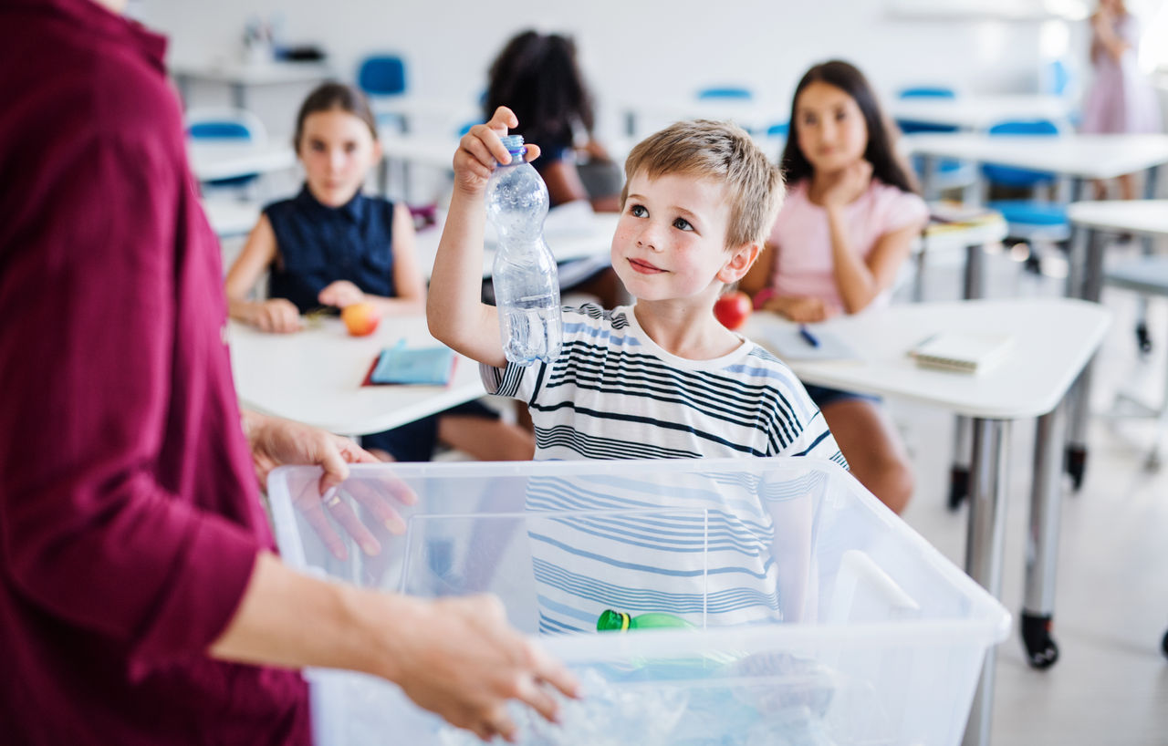 A boy upfront of a classroom with his teacher placing a plastic drink bottle into a plastic bin with other recyclable waste.