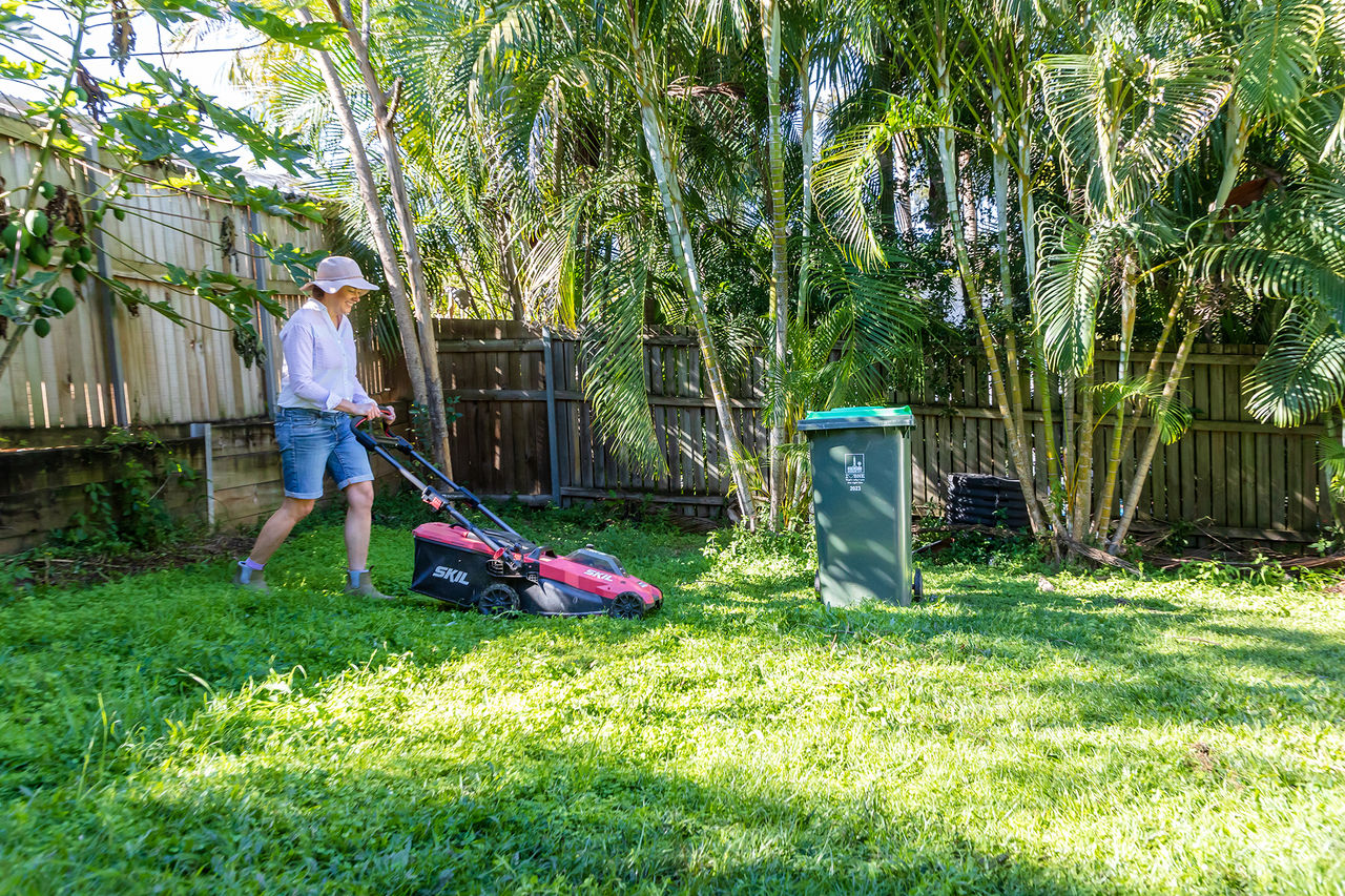 Woman mowing backyard with a Skil lawnmower. Green bin in yard.