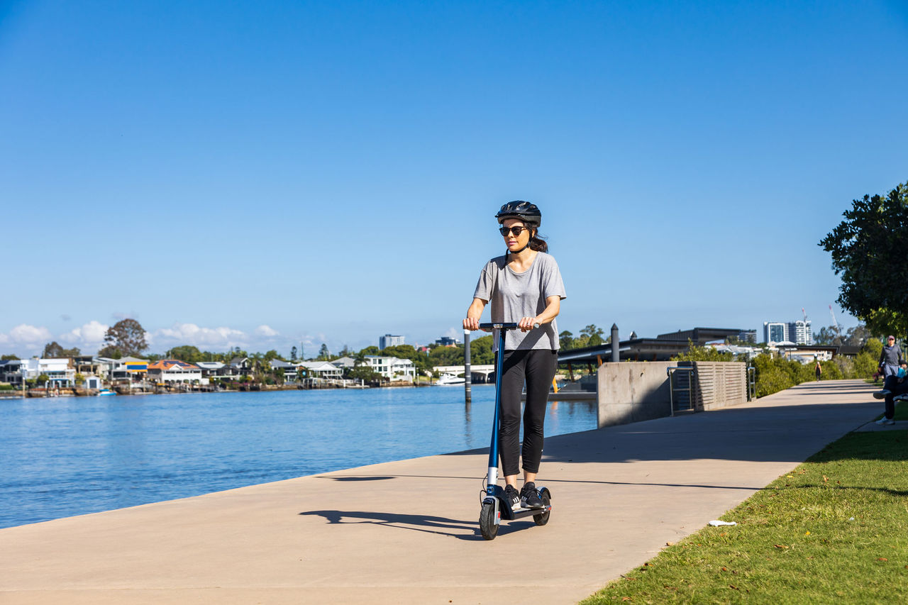 Woman riding an e-scooter along the Brisbane River at New Farm. Bright blue sky as background.