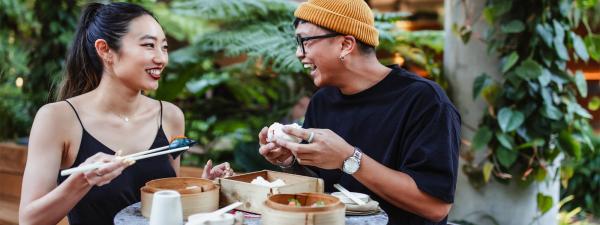 Asian man and woman seated at a table eating a meal with trees in the background